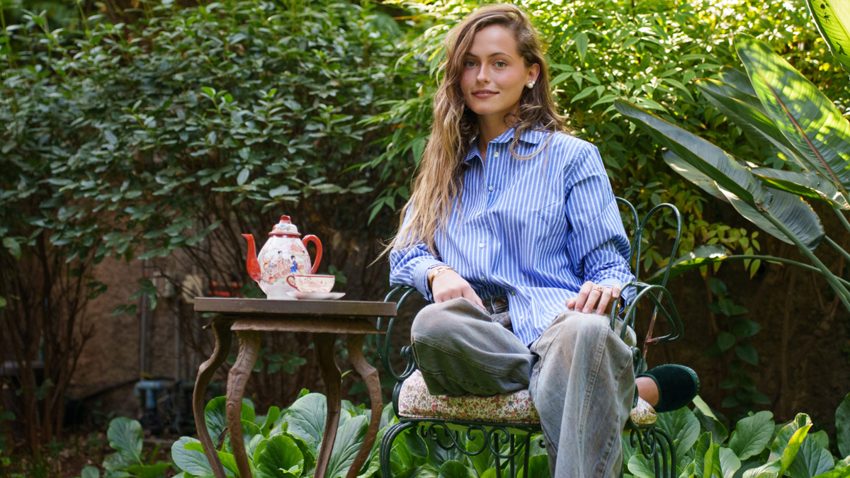 Mujer con camisa de rayas azules sentada en una silla de fierro en un jardín, junto a una tetera pintada a mano